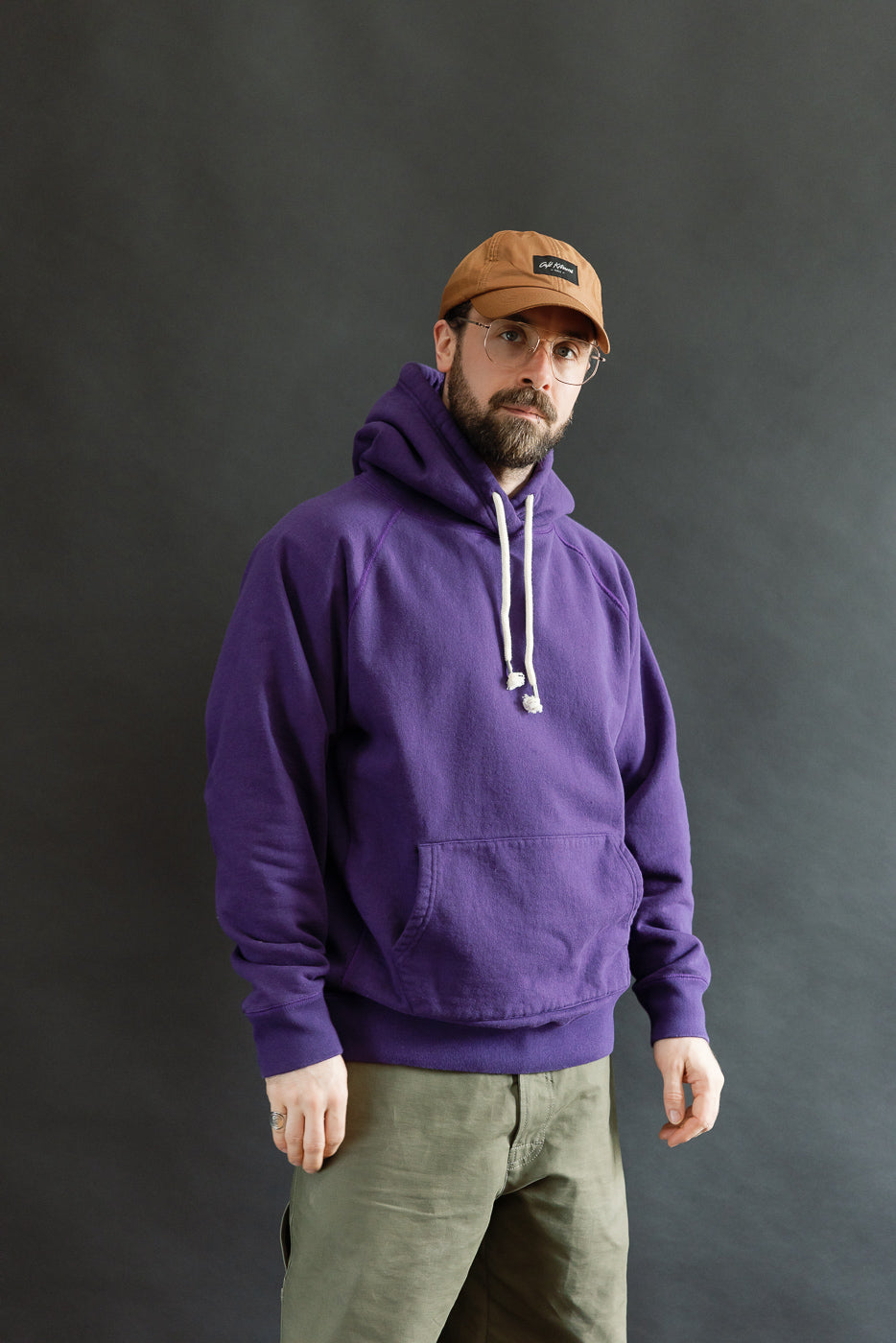 Man wearing Pullover Hoodie - Super Looper French Terry - Purple, showcasing its smooth exterior and textured interior. Styled with olive-green pants and a tan cap, reflecting Dant's premium menswear aesthetic.