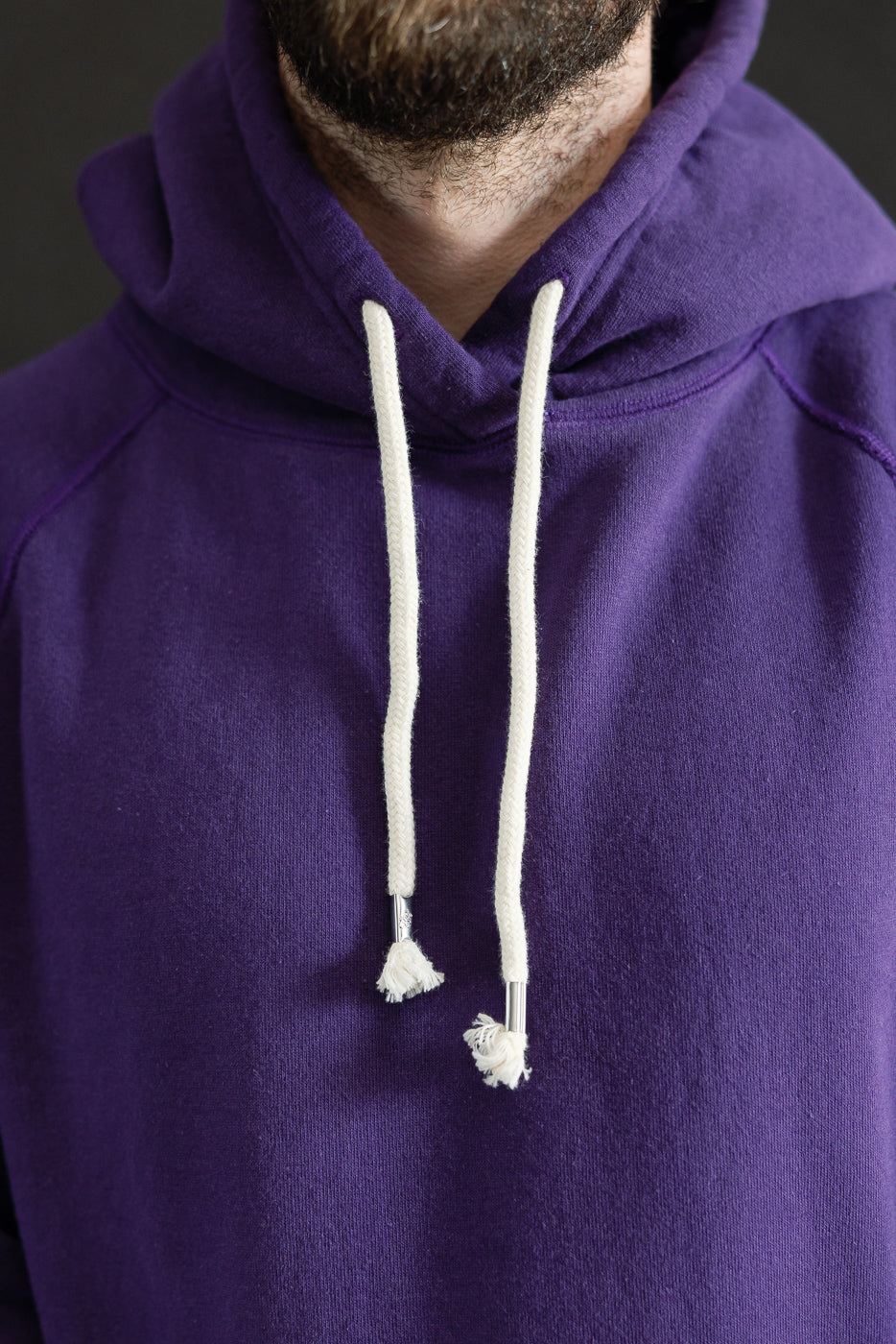 Bearded individual wearing the Pullover Hoodie - Super Looper French Terry - Purple, showcasing the rich texture and classic design with a drawstring hood and visible white drawstrings against a neutral backdrop.
