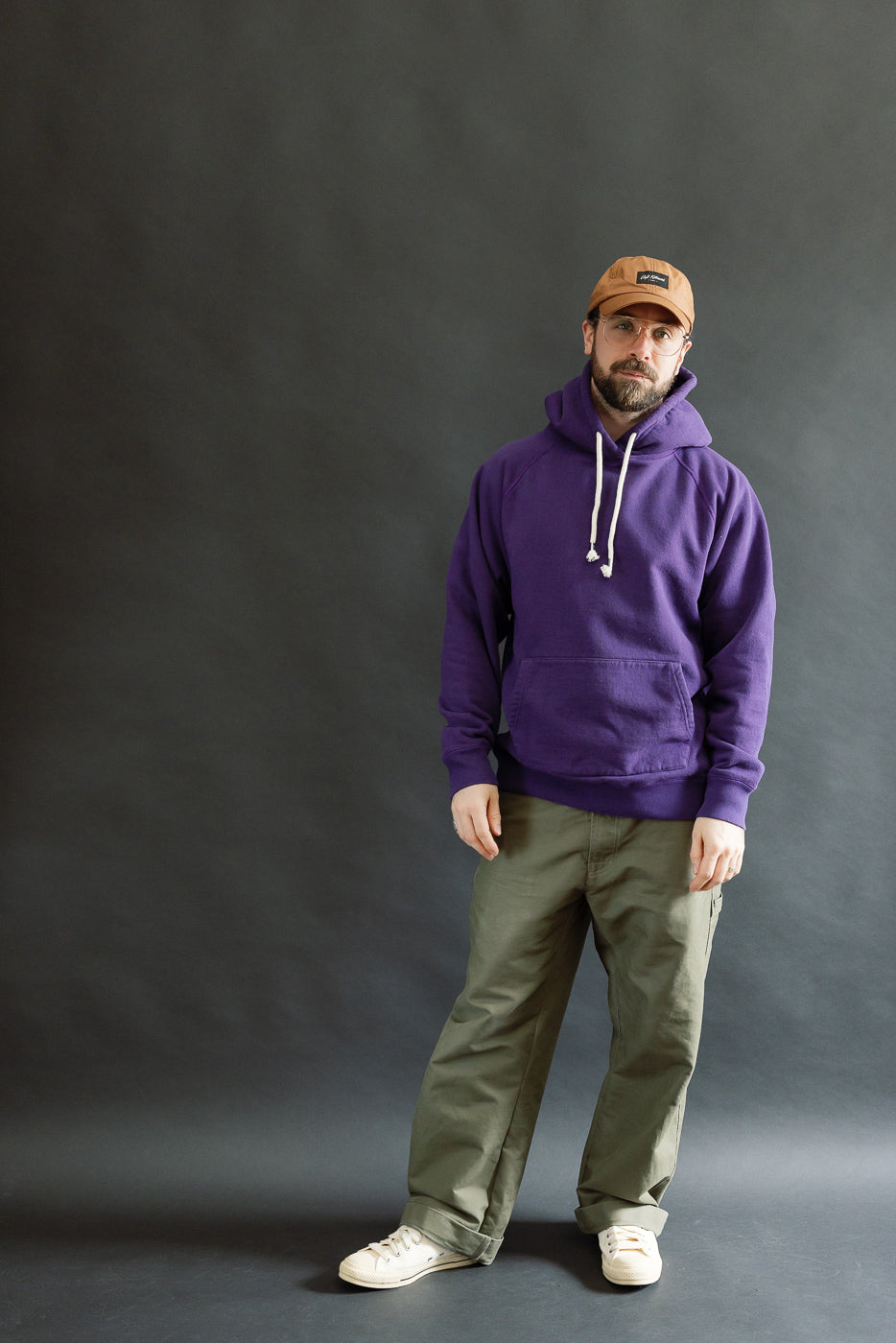 Man wearing the Pullover Hoodie - Super Looper French Terry - Purple, featuring white drawstrings, paired with olive-green trousers and off-white sneakers, standing against a plain dark-gray backdrop.