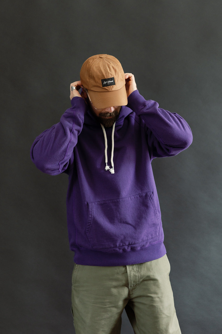 A person adjusts their light brown cap, wearing the Super Looper French Terry Pullover Hoodie in purple, showcasing its smooth exterior and classic design features like raglan sleeves and a kangaroo pocket.