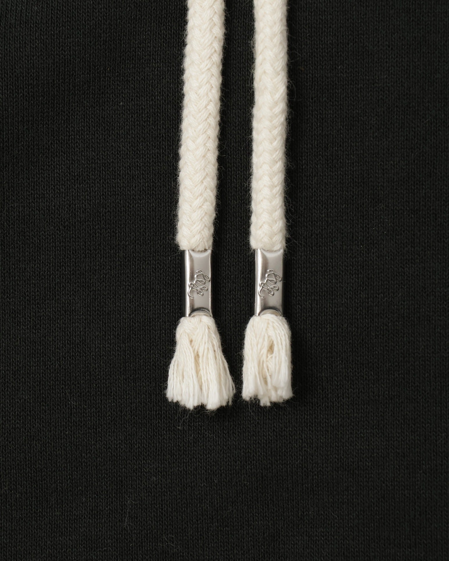 Close-up of pullover hoodie front showing braided cream drawstrings with metal tips on Sumi black French terry fabric.