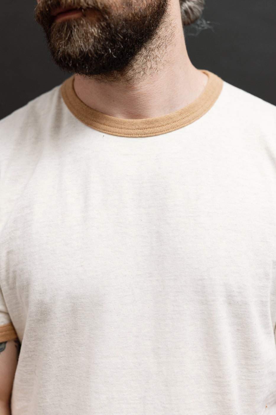 Close-up front view of the ribbed tan crewneck collar on a cream cotton t-shirt.