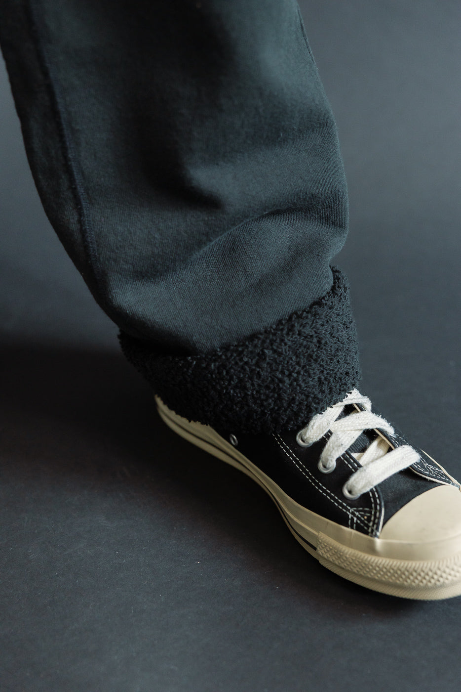 Close-up of male model's lower leg showing black selvedge jeans with fuzzy hem cuff, paired with black low-top sneakers