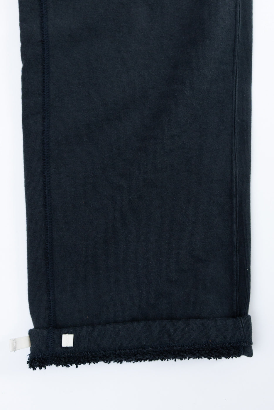 Close-up of black sweatpants leg, front view, showing raw hem, side seam, and small white tag detail.