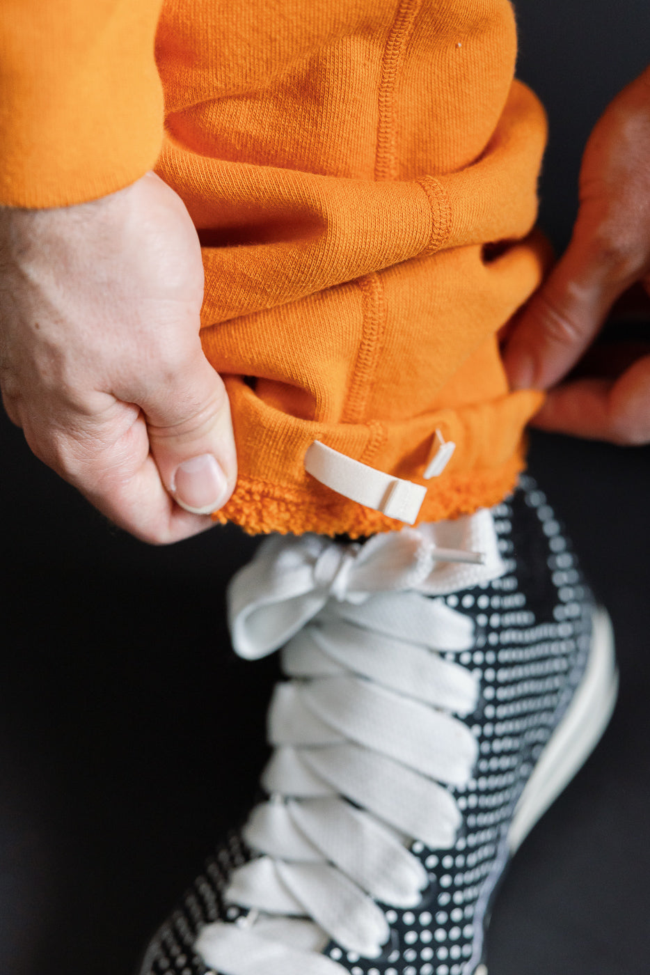 Close-up of male model's orange hoodie hem with white drawstring toggle, showcasing ribbed texture and fit over black-and-white sneaker