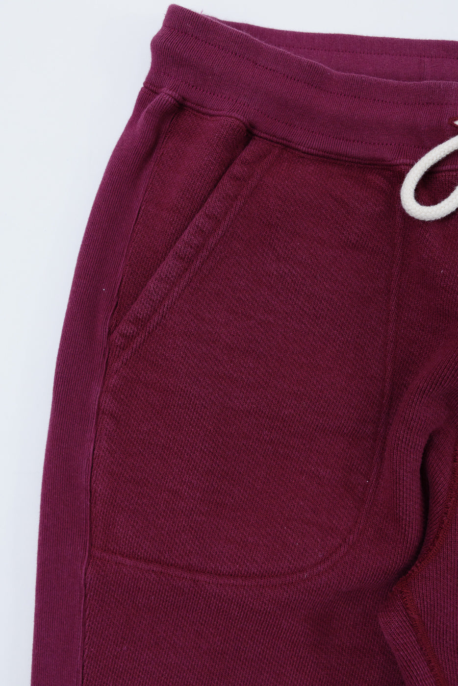 Close-up of the front left pocket and waistband of burgundy corduroy trousers with white drawstring.