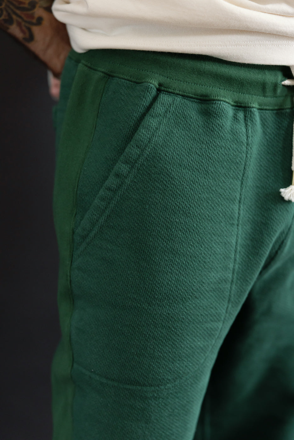 Close-up of the left side pocket and ribbed waistband of green cotton sweatpants worn by a male model.