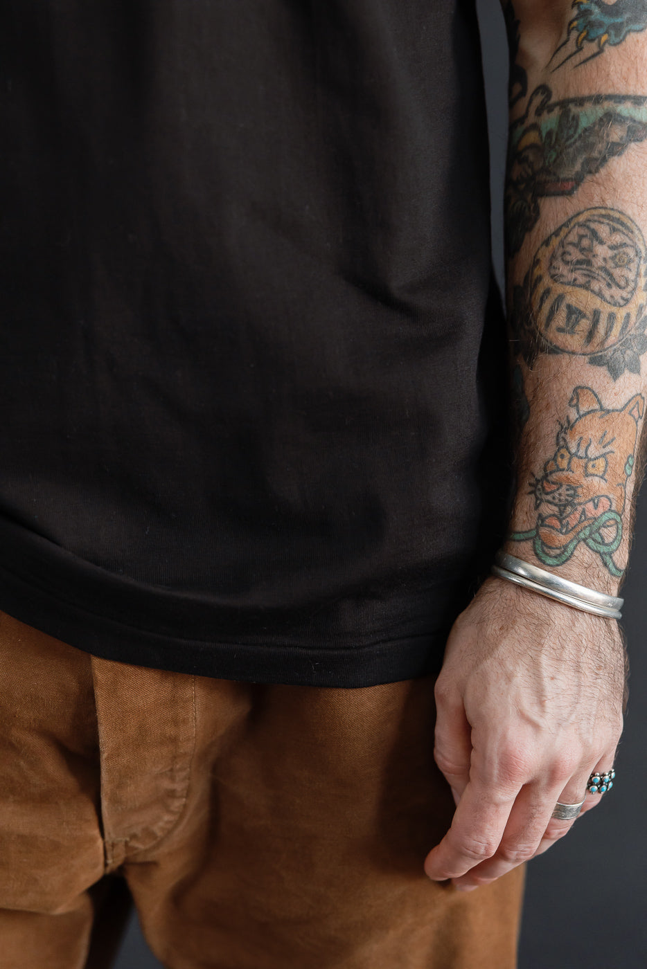 Close-up of the front of a male model's black t-shirt and brown trousers, showing arm tattoos.