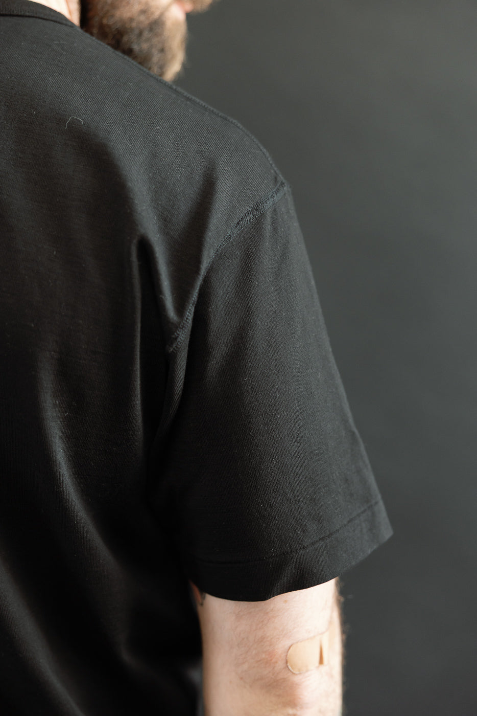 Person wearing the Ultimate Pima Tsuriami Crewneck T-Shirt - Black, showcasing its smooth fabric and comfortable fit, highlighted by a small bandage on the right tricep against a neutral background.