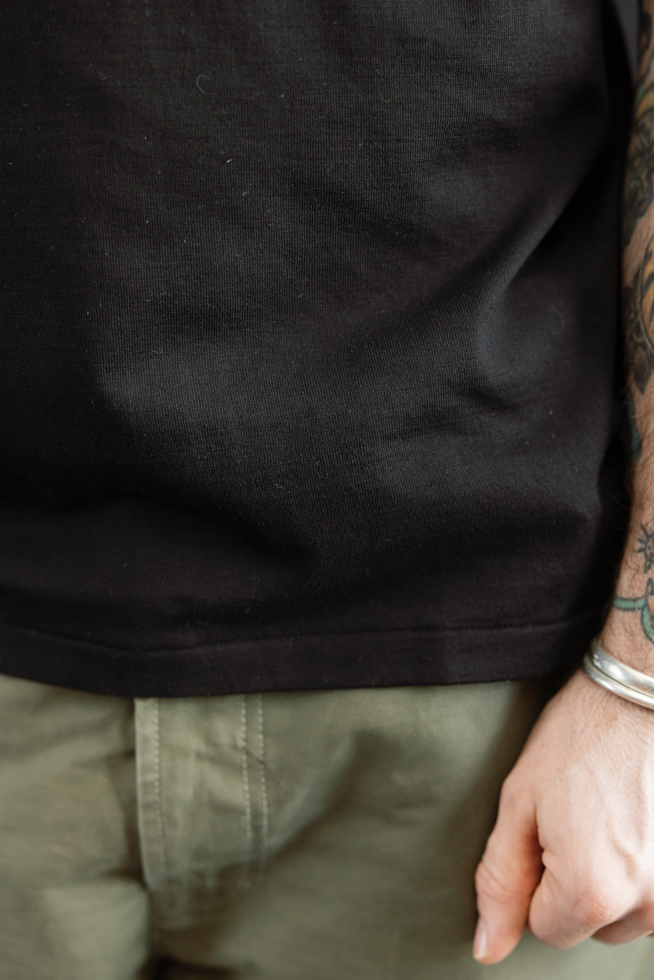 Close-up of the Ultimate Pima Tsuriami Crewneck T-Shirt - Black, showcasing its smooth texture and lightweight fabric over olive-green pants, complemented by tattooed forearm with silver bangles.