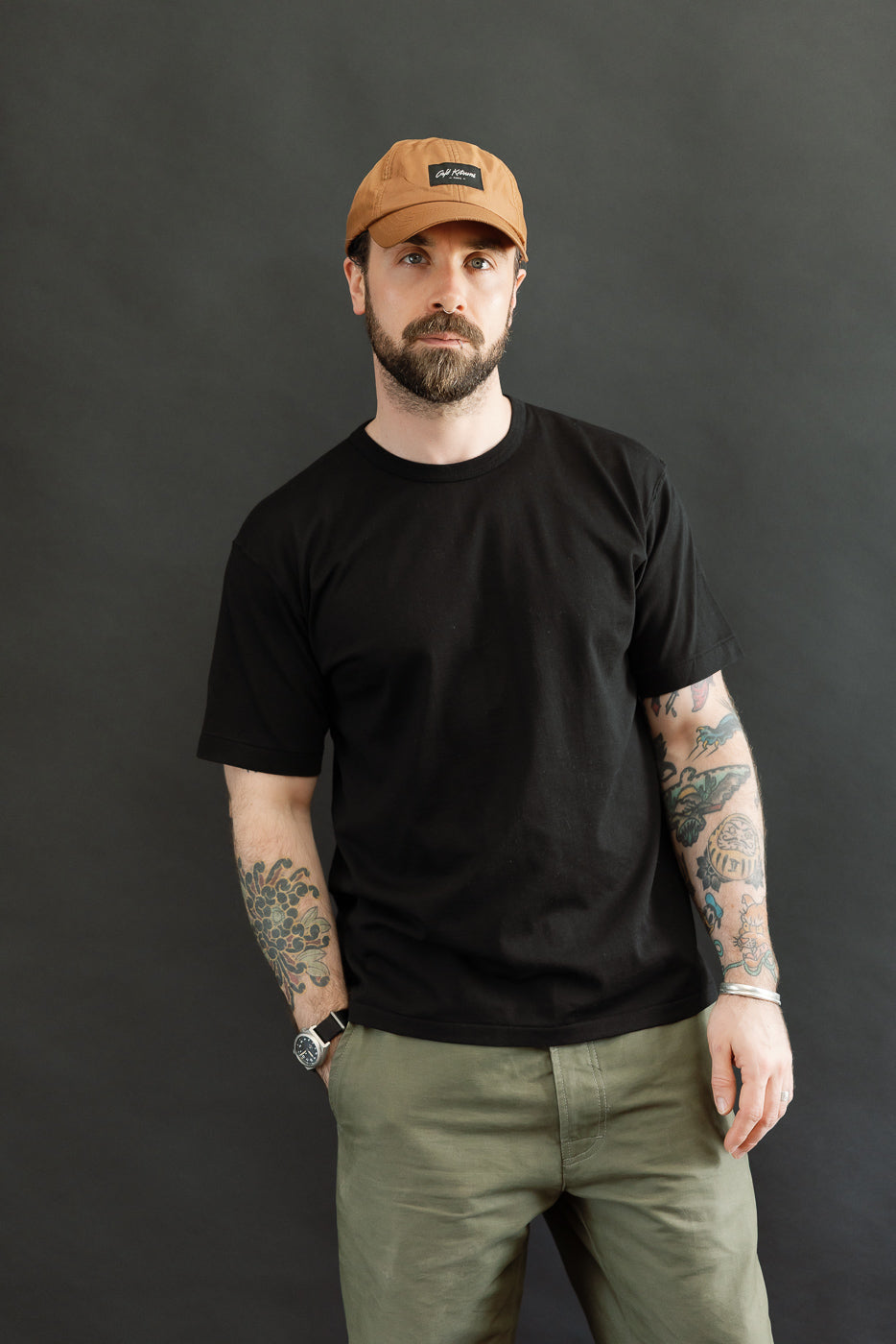 Man wearing an Ultimate Pima Tsuriami Crewneck T-Shirt - Black, styled with olive-green trousers and a light-brown baseball cap, showcasing colorful tattooed forearms against a dark-gray backdrop.