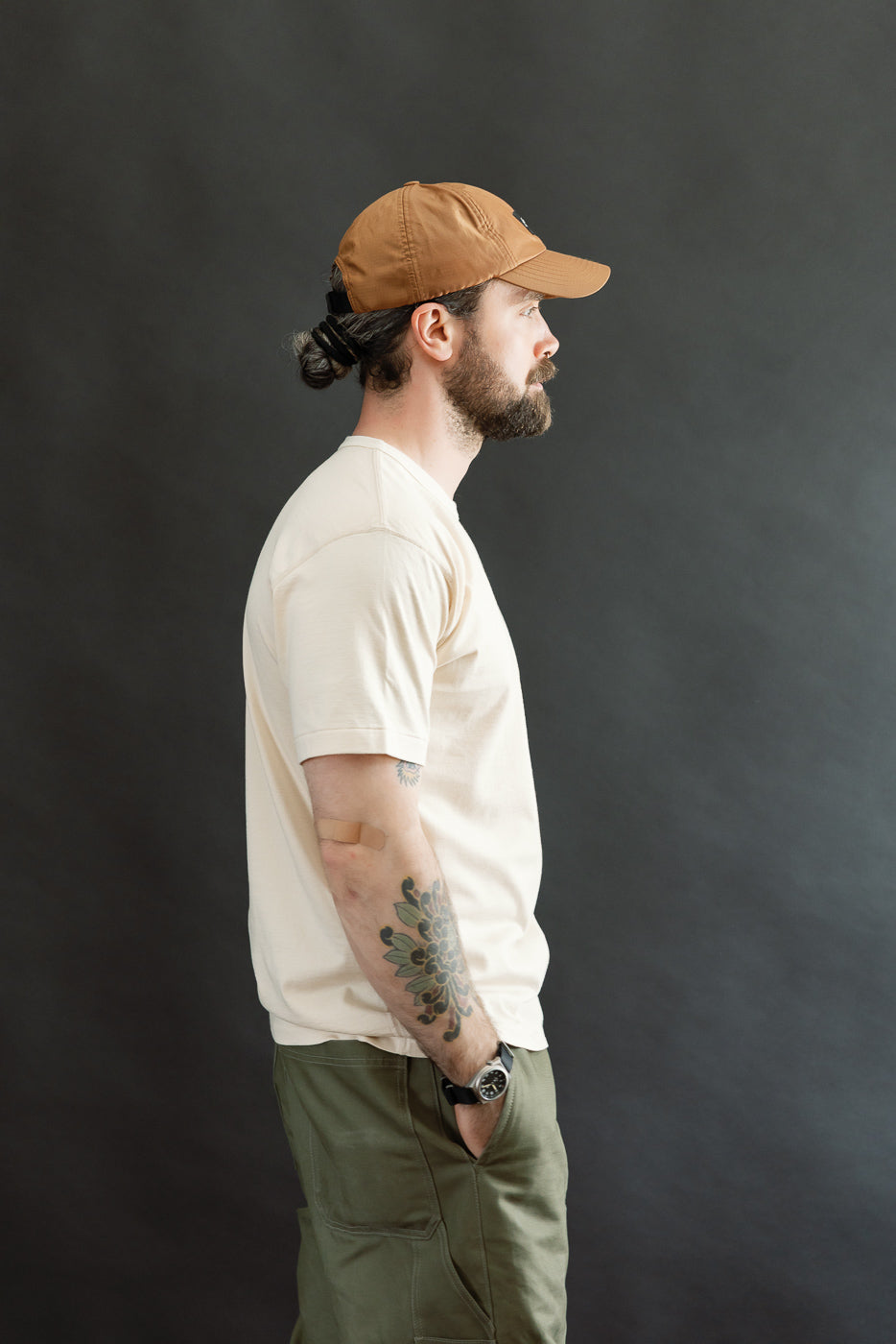 A bearded man in profile wearing the Ultimate Pima Tsuriami Crewneck T-Shirt in Ecru, showcasing its smooth texture, light weight, and athletic fit, styled with olive-green pants and a brown cap.