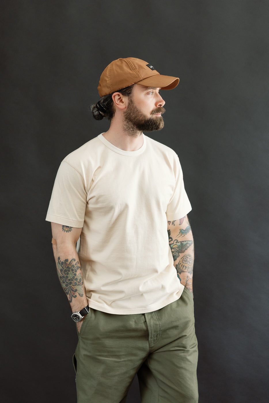 A man models the Ultimate Pima Tsuriami Crewneck T-Shirt - Ecru, showcasing its lightweight, smooth texture and athletic fit, paired with olive-green pants and a brown baseball cap.