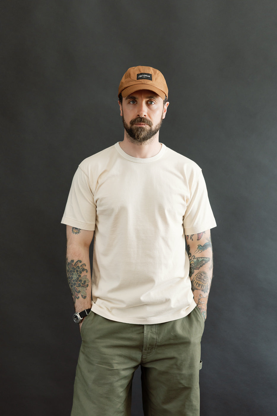 Ultimate Pima Tsuriami Crewneck T-Shirt - Ecru, worn by a bearded man with tattoos, features a light-beige, smooth organic cotton fabric, paired with olive-green pants and a brown cap, embodying Dant's premium menswear ethos.