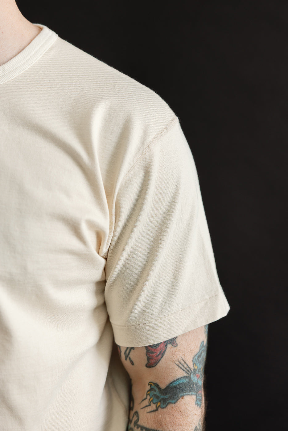 Close-up of the Ultimate Pima Tsuriami Crewneck T-Shirt - Ecru, highlighting its soft, light-beige fabric and short-sleeve design, paired with a tattooed forearm showcasing a stylized panther motif.