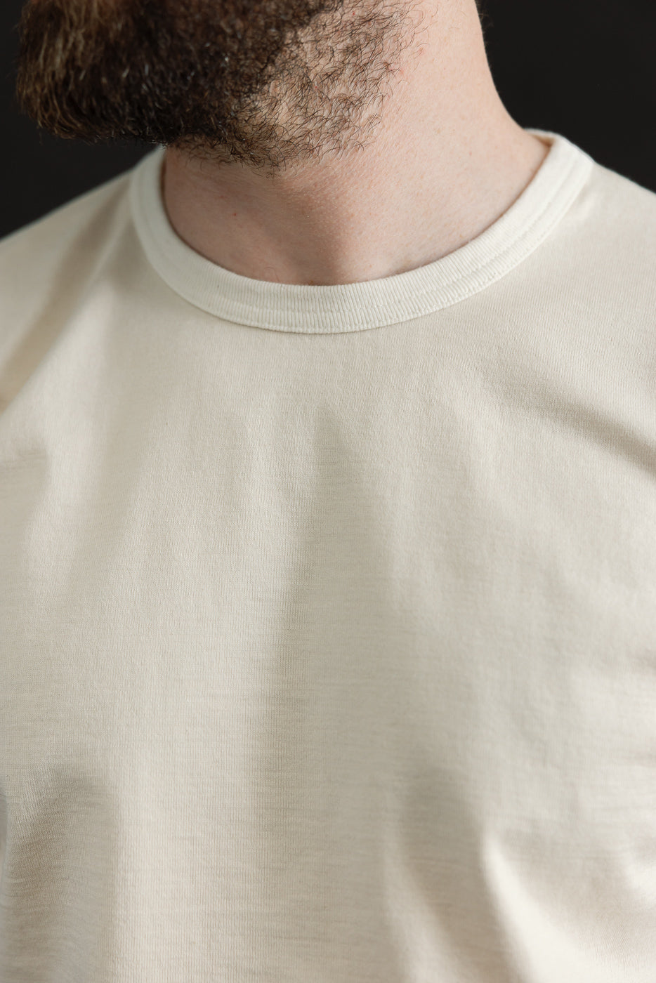 Close-up of a person wearing the Ultimate Pima Tsuriami Crewneck T-Shirt - Ecru, highlighting its smooth texture and light-cream color against a dark background, emphasizing the premium fabric's softness.