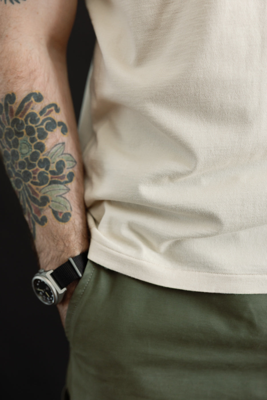 Model wearing Ultimate Pima Tsuriami Crewneck T-Shirt - Ecru, showcasing the smooth, lightweight fabric with a floral tattoo visible on the forearm, standing with a hand in olive-green pants pocket.