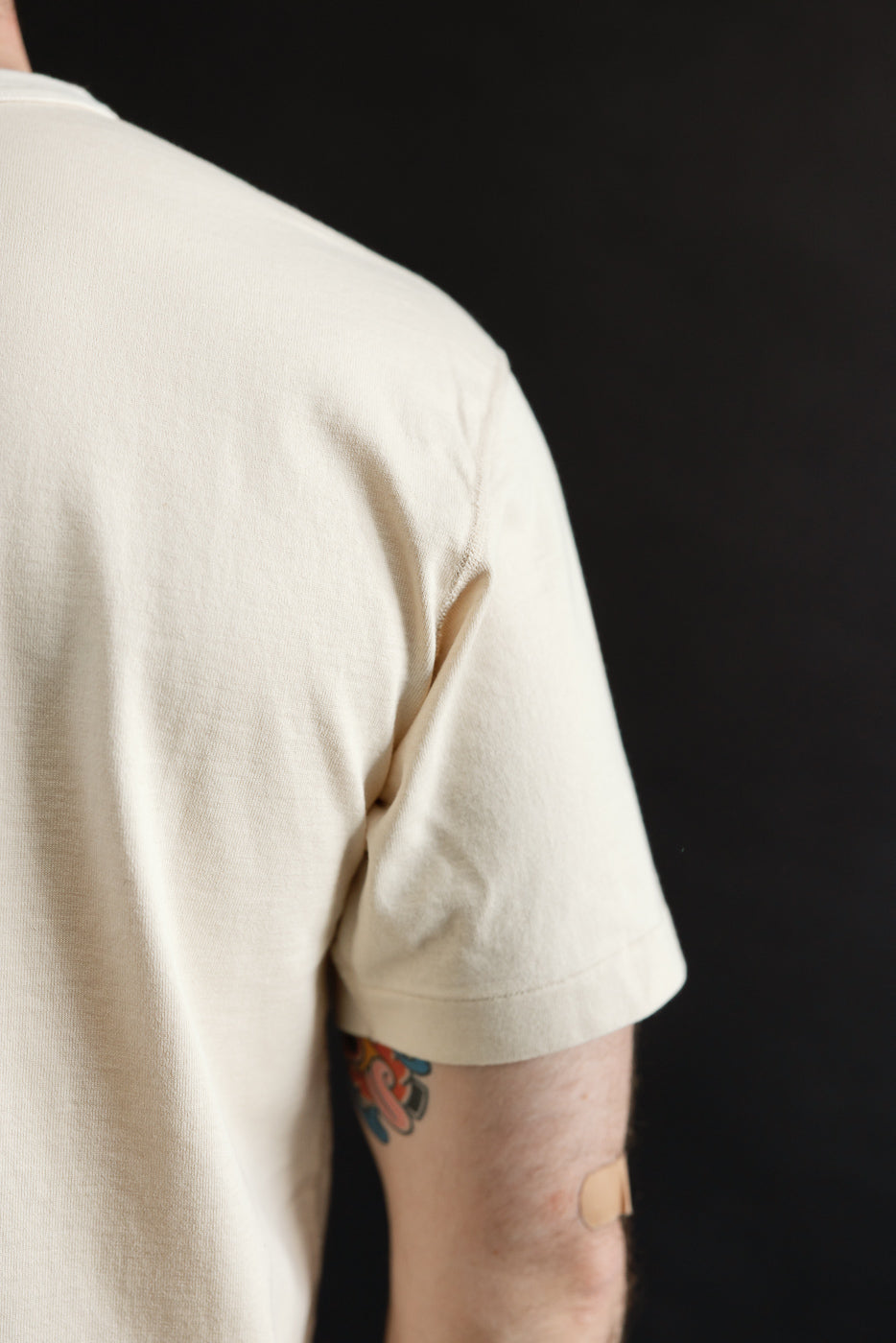 Person's back and upper arm in a light-beige Ultimate Pima Tsuriami Crewneck T-Shirt – Ecru, showcasing a smooth finish and visible tattoo with bandage, reflecting premium craftsmanship.