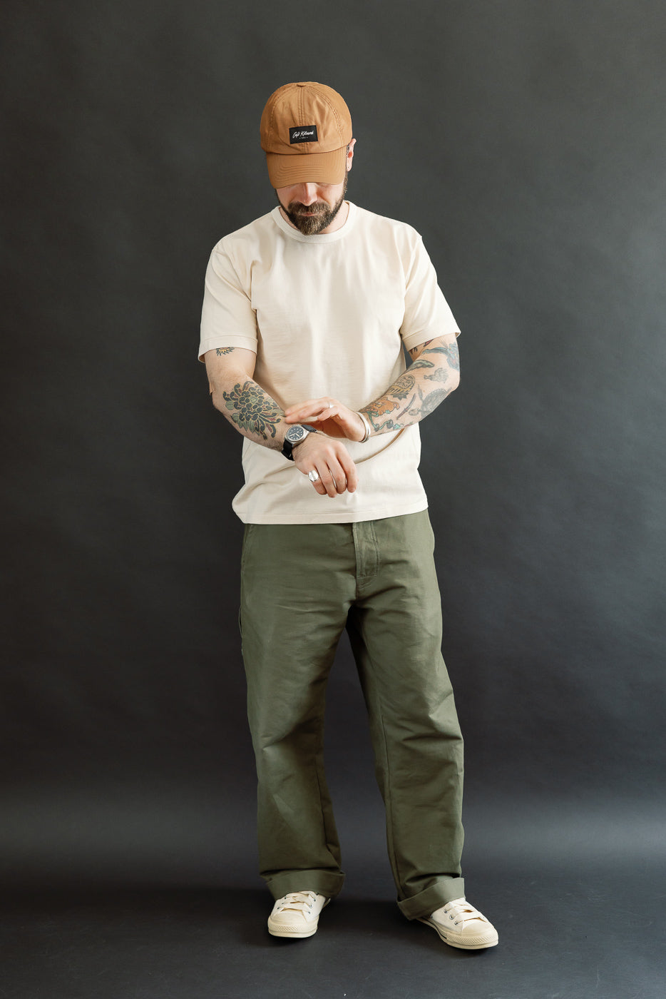 A bearded man models the Ultimate Pima Tsuriami Crewneck T-Shirt in ecru, paired with olive pants and canvas sneakers, highlighting its lightweight, smooth texture and athletic fit against a dark-gray backdrop.