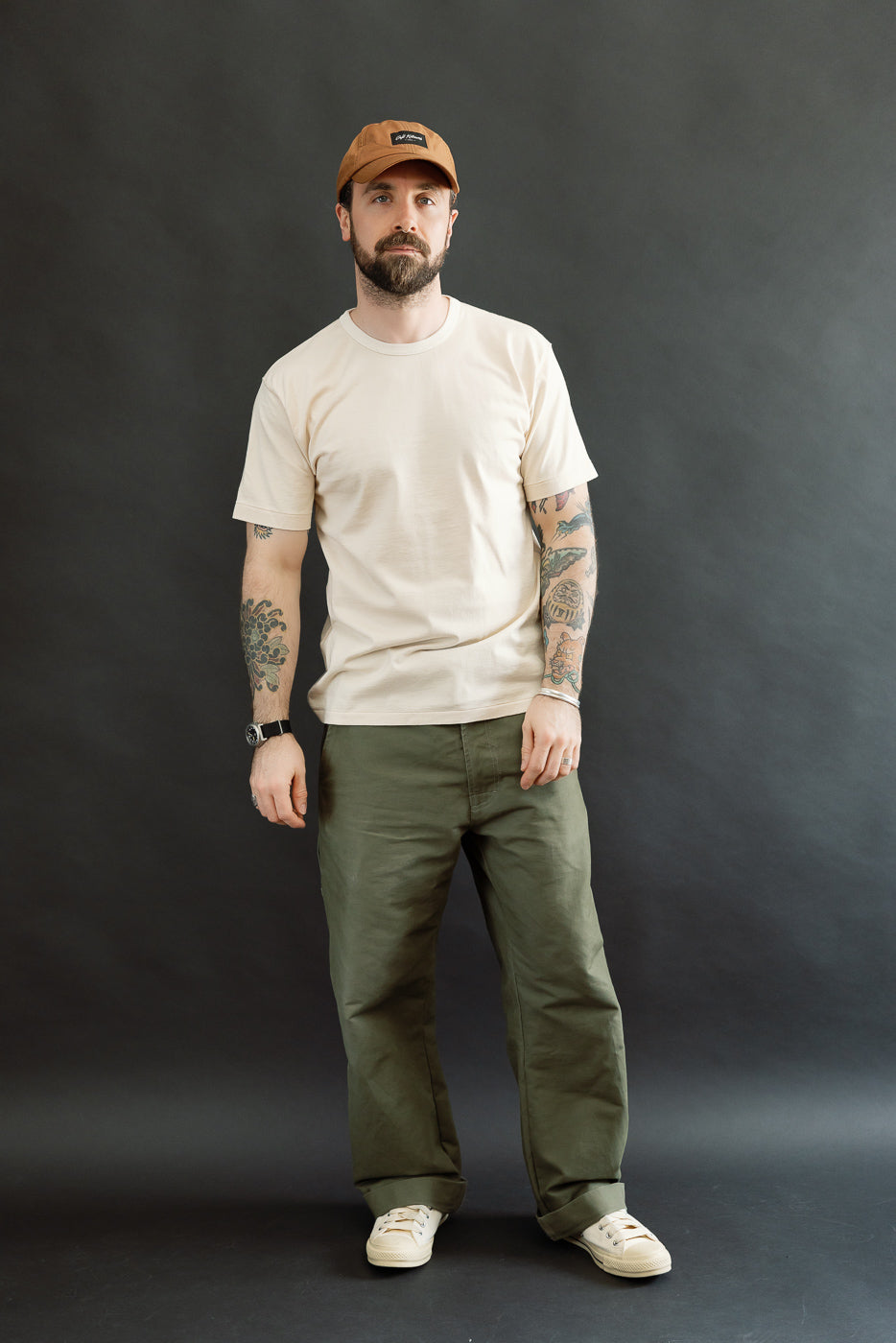 Full-body shot of a bearded Caucasian man wearing the Ultimate Pima Tsuriami Crewneck T-Shirt in Ecru, styled with olive-green pants and off-white sneakers, showcasing the relaxed, premium menswear look.