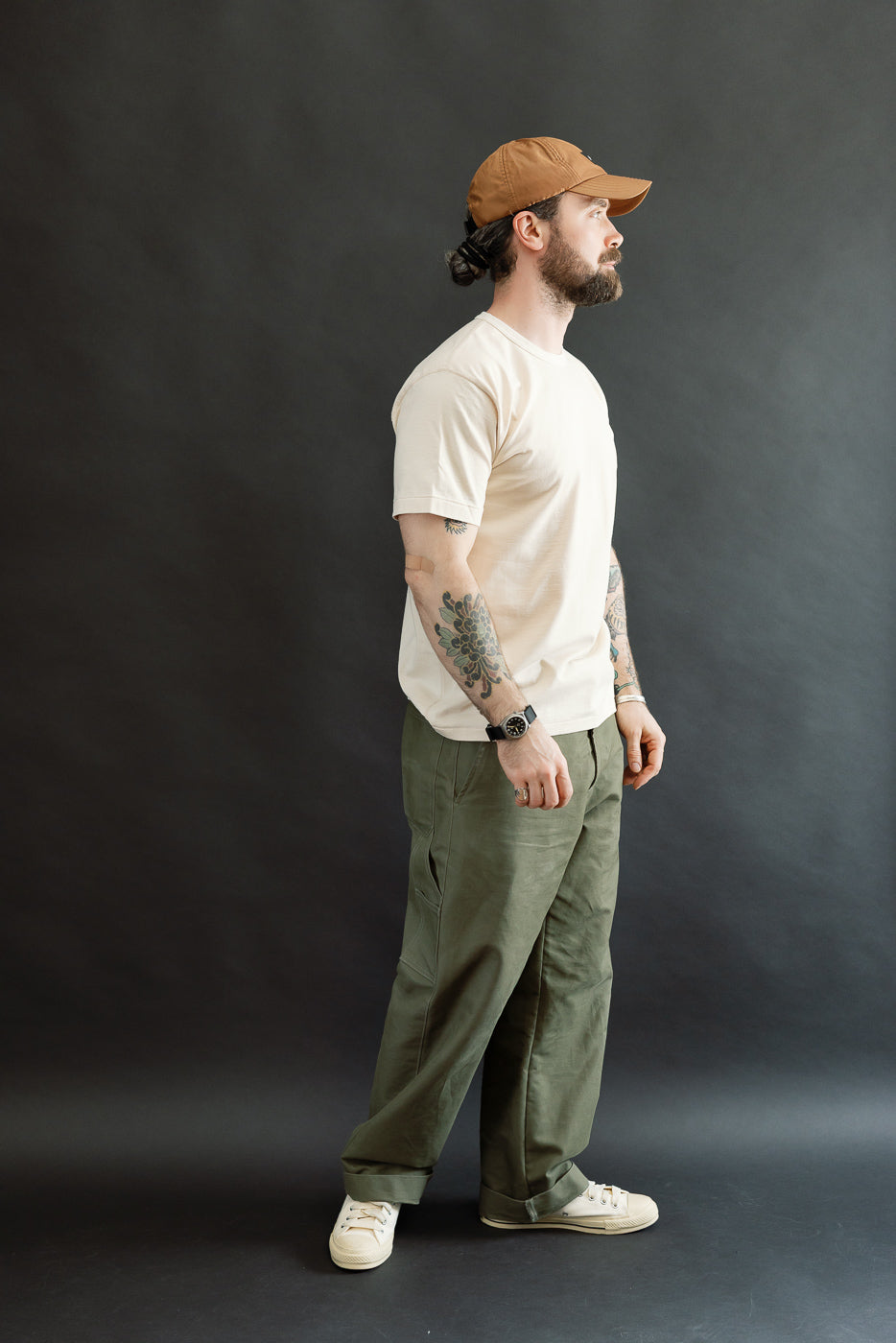 A bearded man models the Ultimate Pima Tsuriami Crewneck T-Shirt - Ecru, showcasing its lightweight, smooth fabric and athletic fit, paired with olive-green pants and off-white sneakers.