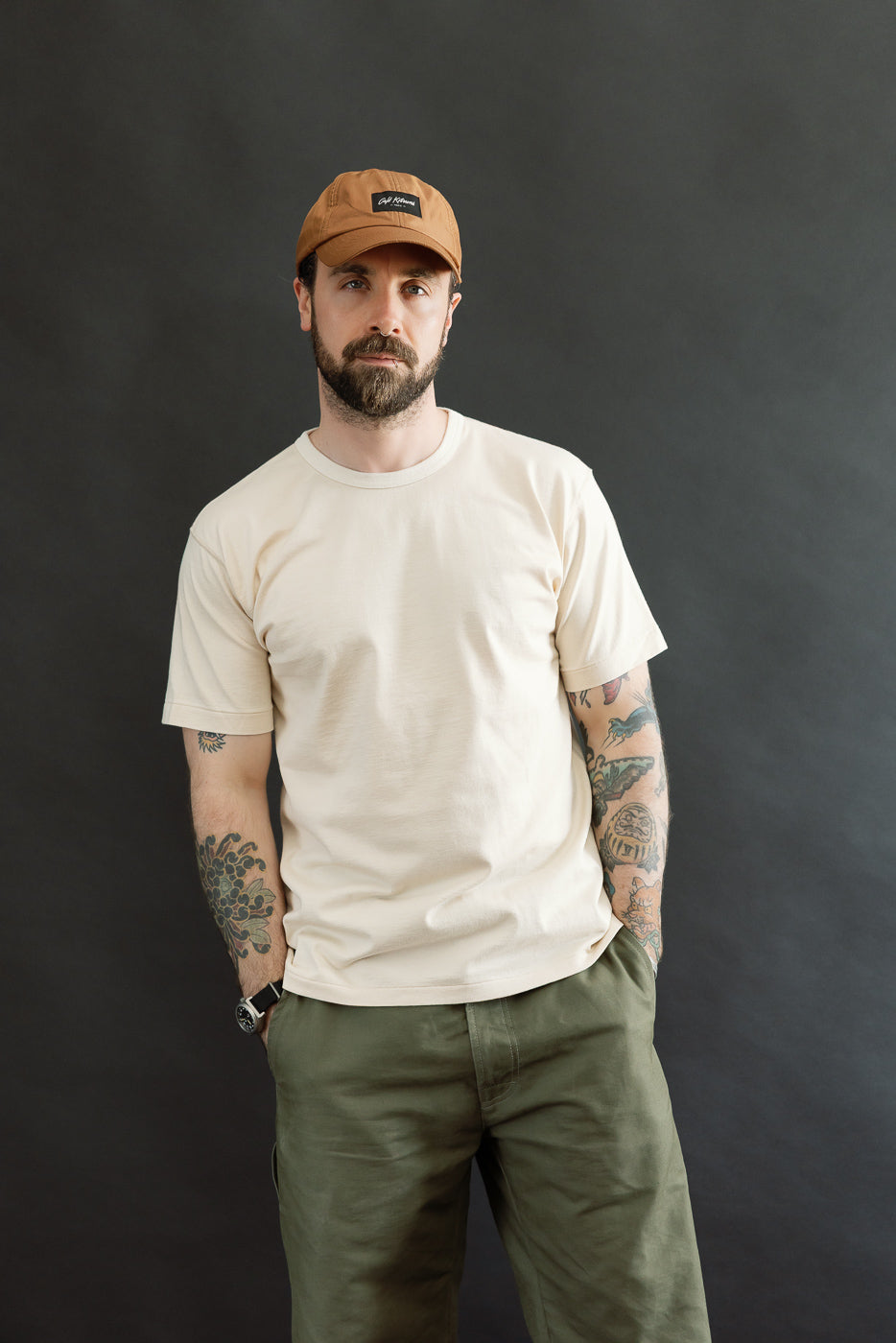 Man wearing the Ultimate Pima Tsuriami Crewneck T-Shirt - Ecru, showcasing its lightweight, smooth organic Pima cotton design, paired with olive-green pants and a tan baseball cap, embodying premium menswear style.