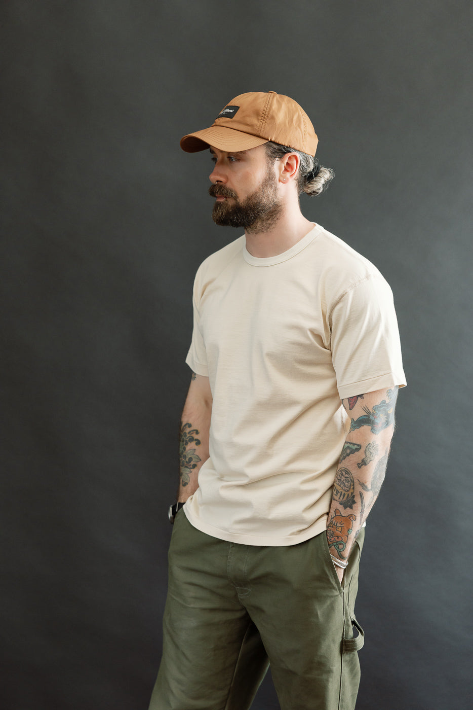 Man wearing Ultimate Pima Tsuriami Crewneck T-Shirt - Ecru, showcasing its light, smooth texture and athletic fit. He's styled in olive-green pants and a tan cap, embodying Dant's heritage-inspired fashion ethos.
