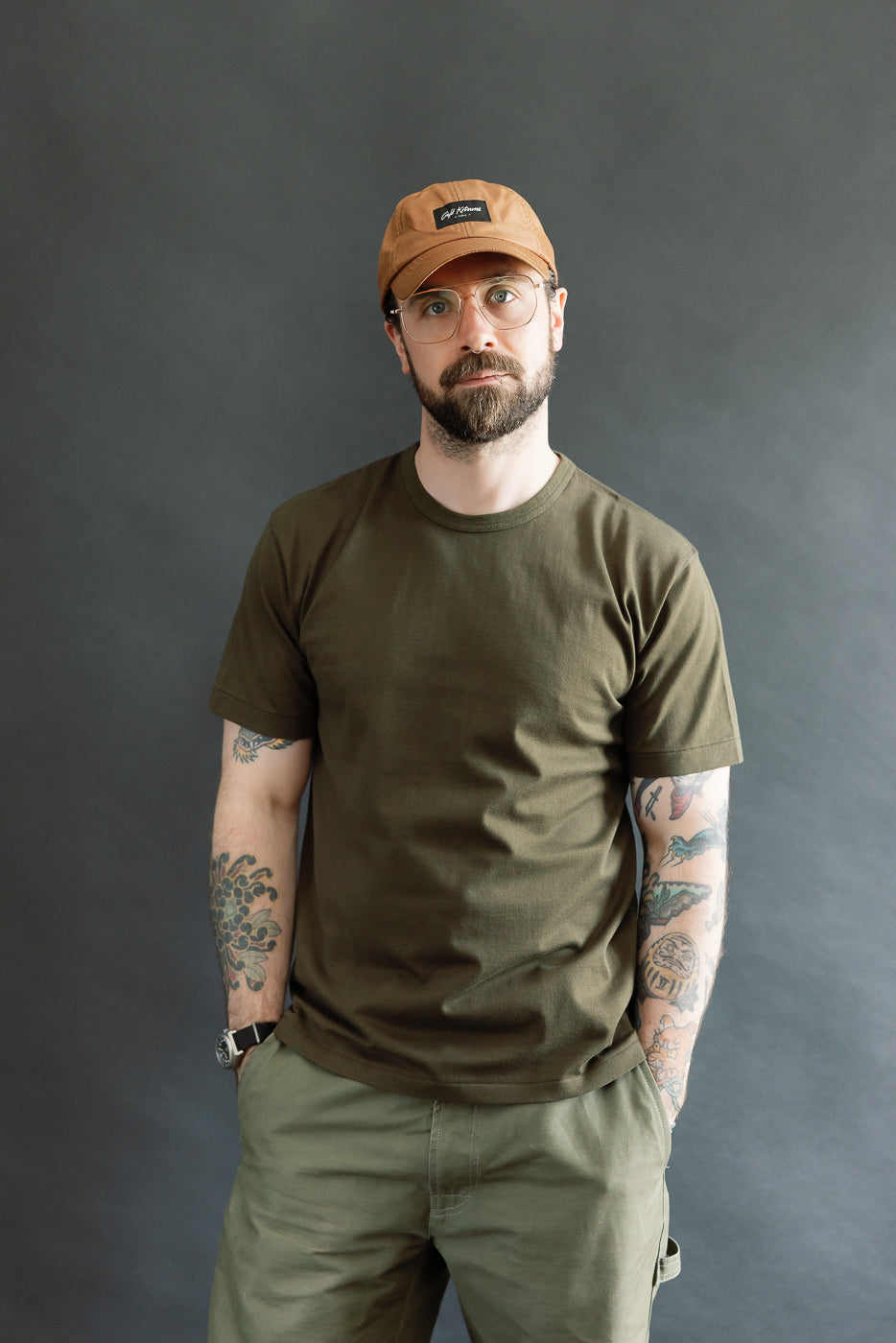 A man models the Ultimate Pima Tsuriami Crewneck T-Shirt - Khaki, showcasing its soft, smooth texture and athletic fit, complemented by tattoos and a light brown baseball cap.