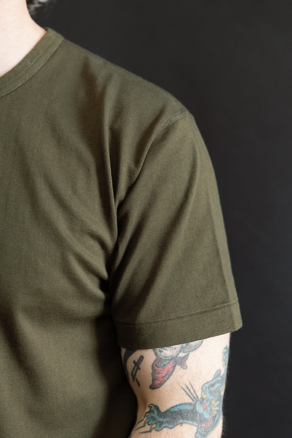 Close-up of a model wearing the Ultimate Pima Tsuriami Crewneck T-Shirt - Khaki, showcasing its smooth texture and lightweight fabric. The tattooed forearm adds a touch of style against the dark background.