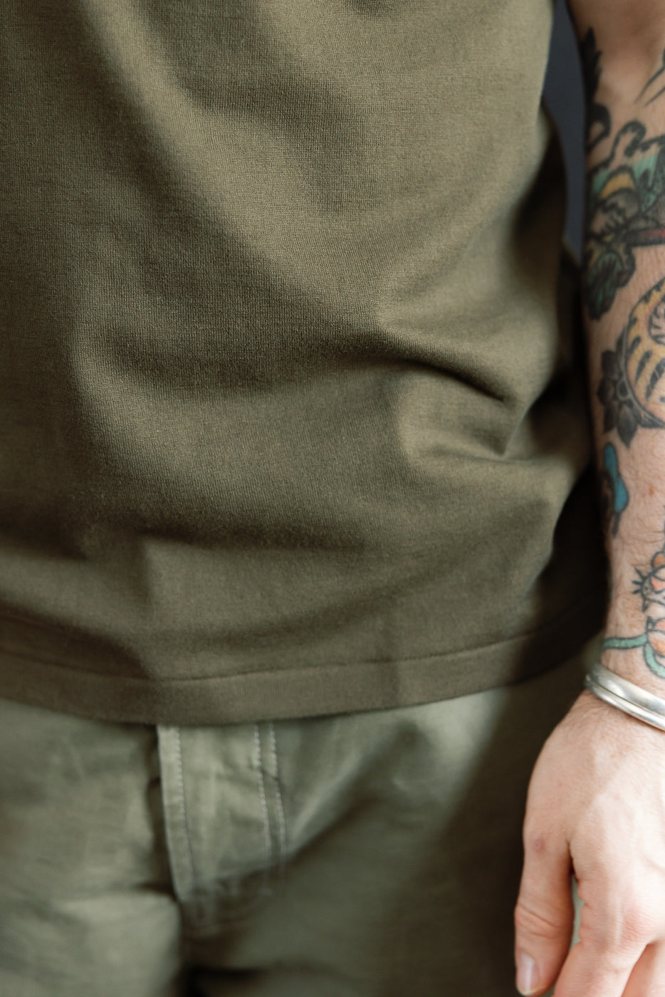 A person's midsection showcasing the Ultimate Pima Tsuriami Crewneck T-Shirt - Khaki, highlighting its smooth texture and lightweight organic cotton fabric with colorful tattoos and silver bracelets visible on the forearm.
