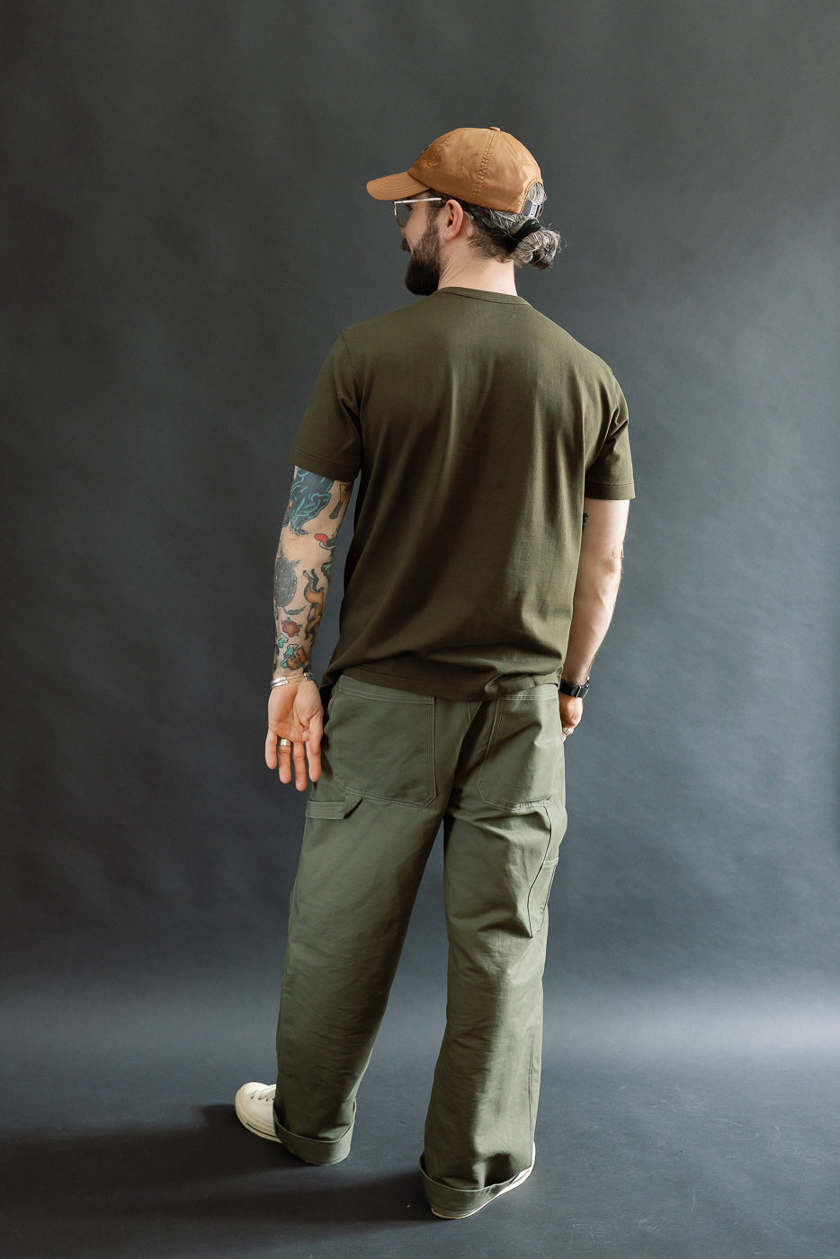 Man in olive-green Ultimate Pima Tsuriami Crewneck T-Shirt and cargo pants, showcasing tattoos and cap, embodies a casual, outdoorsy style.
