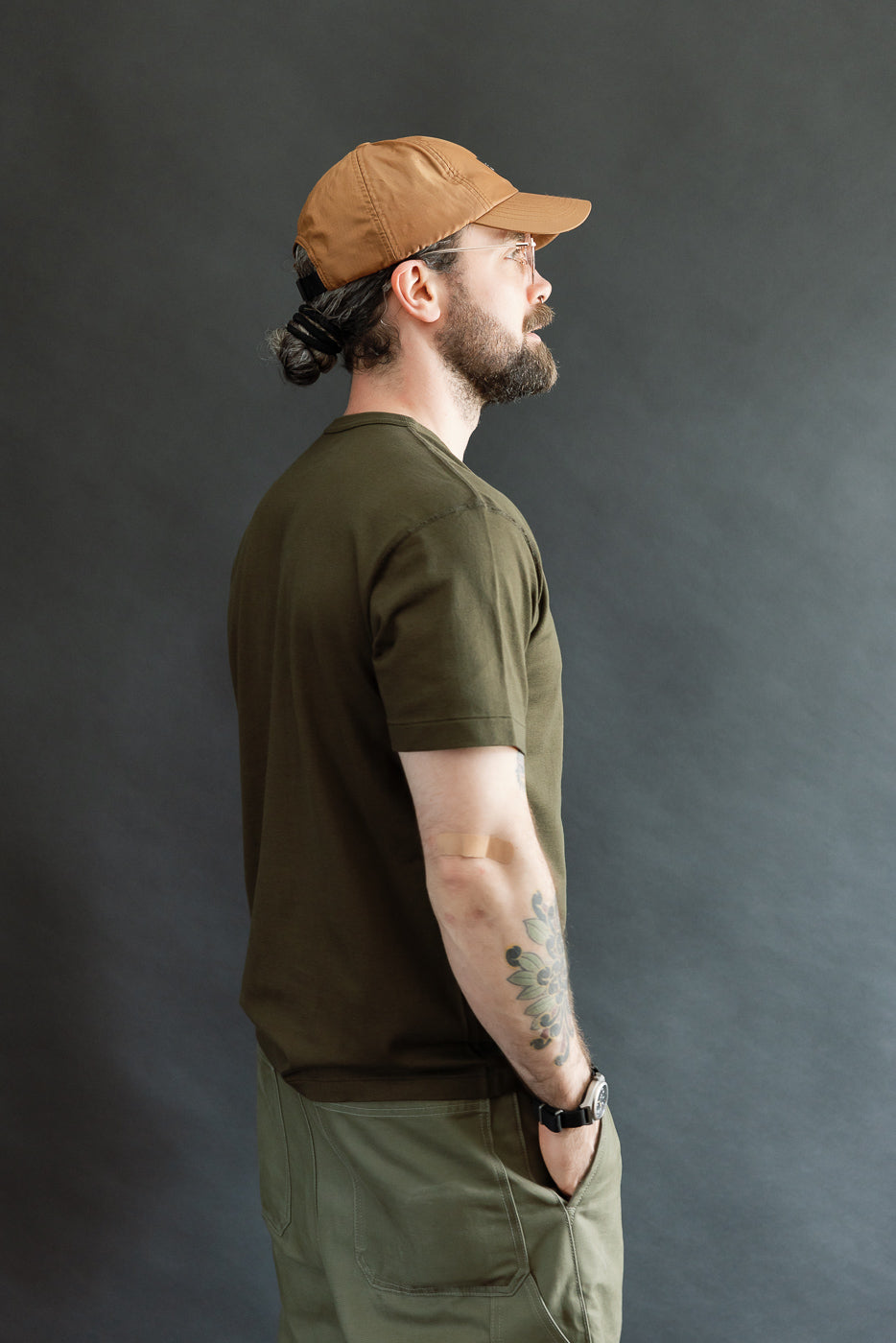 A man in profile wears an Ultimate Pima Tsuriami Crewneck T-Shirt - Khaki, showcasing its smooth, lightweight fabric and athletic fit, paired with a brown cap and olive-green pants.