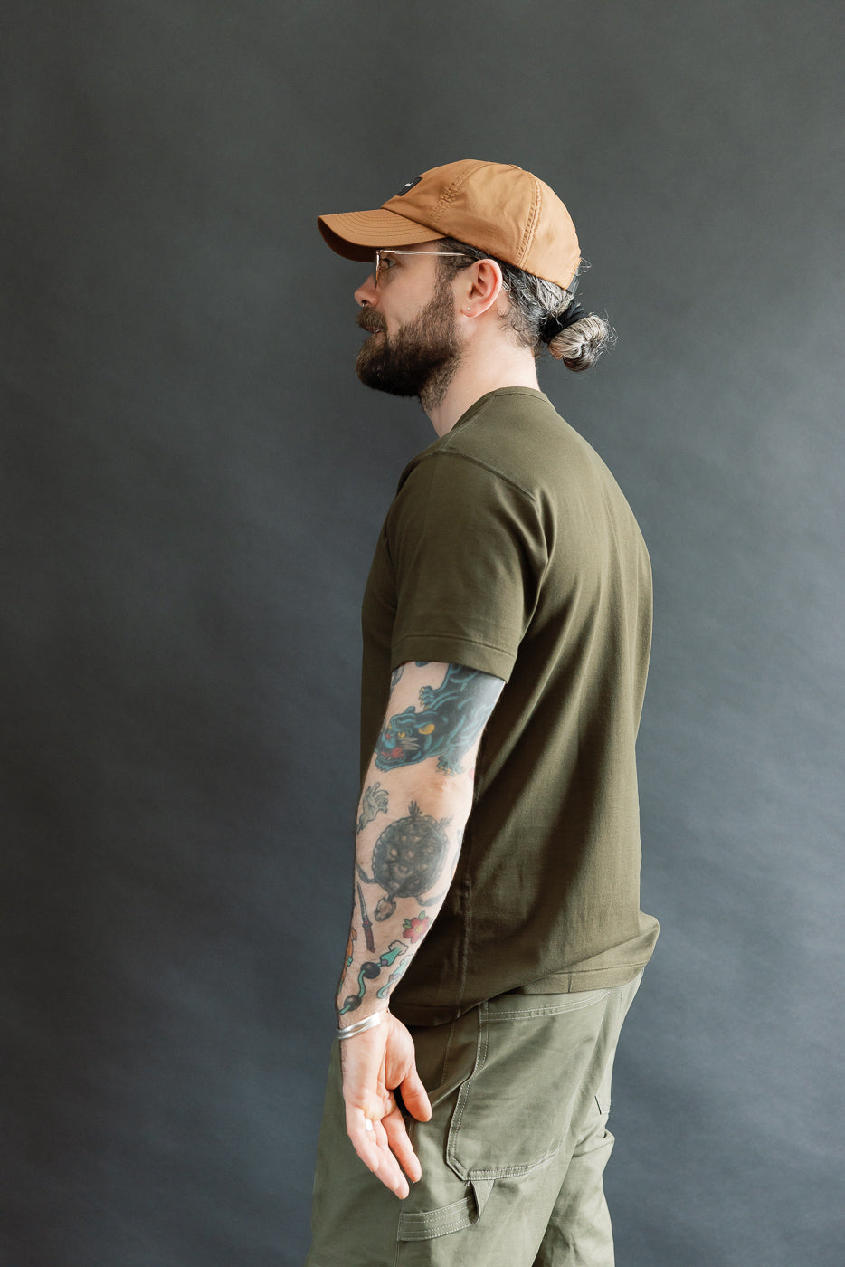 A bearded man in wire-frame glasses wears the Ultimate Pima Tsuriami Crewneck T-Shirt - Khaki, showcasing its smooth, lightweight texture and casual fit against a plain background.