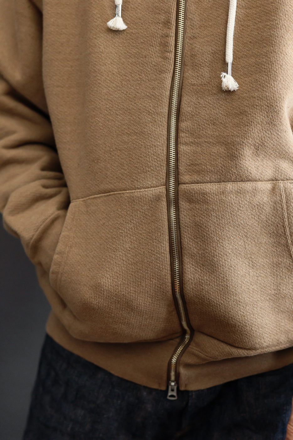 Close-up front view of a tan zip-up hoodie featuring a metal zipper and ribbed fabric texture.