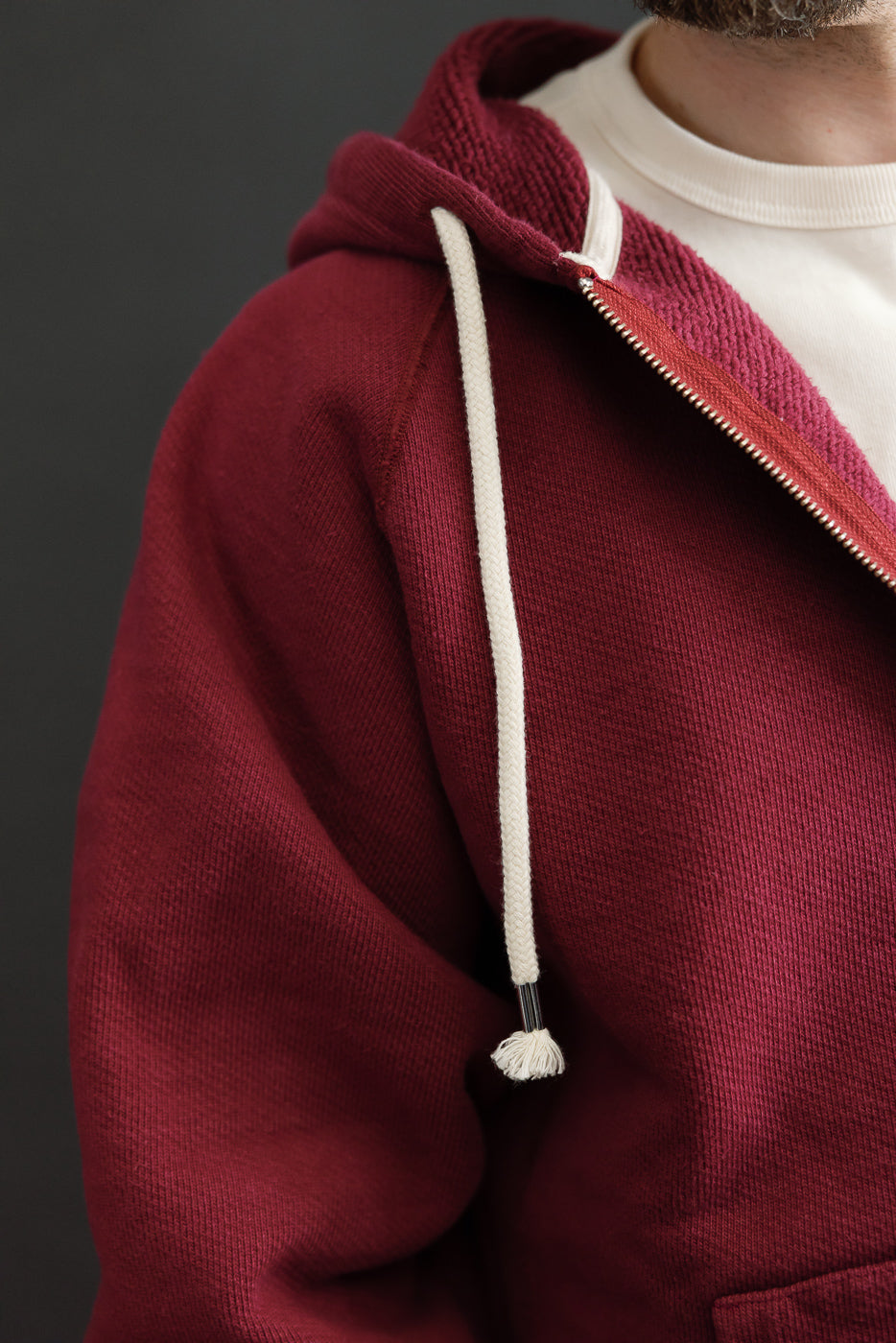 Close-up of the front left side of a burgundy zip-up hoodie showing fabric texture and white drawstring.