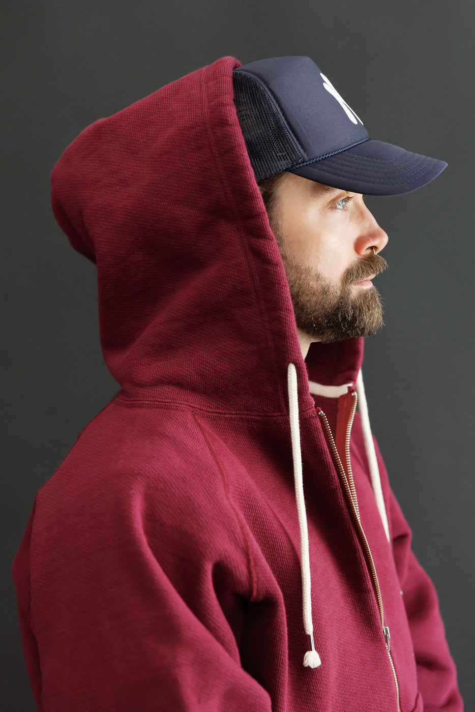 Close-up side profile of a male model wearing a burgundy hooded sweatshirt and a navy blue mesh cap.