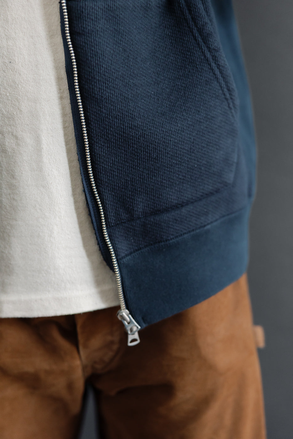 Close-up side view of a navy blue zip-up hoodie's metal zipper and pocket detail on a male model.