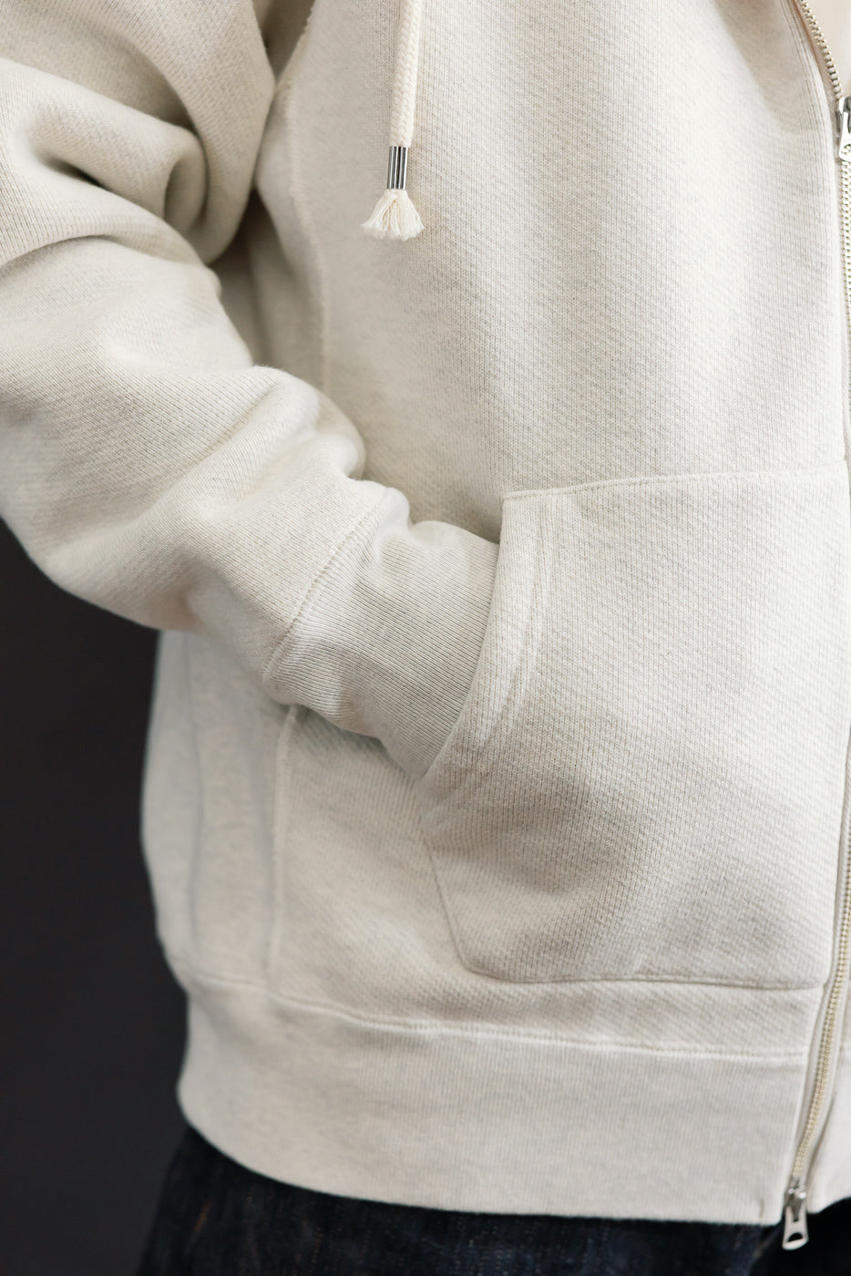 Close-up side view of a male model's cream zip-up hoodie, showing the pocket and ribbed cuff.