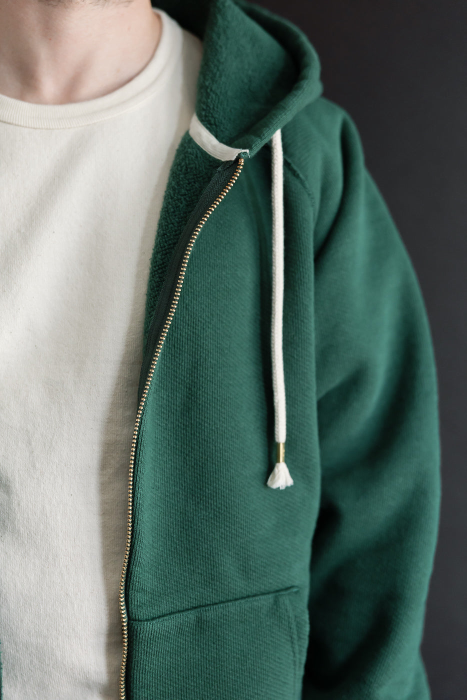 Close-up side view of a green zip-up hoodie with brass zipper and white drawstring on a male model.