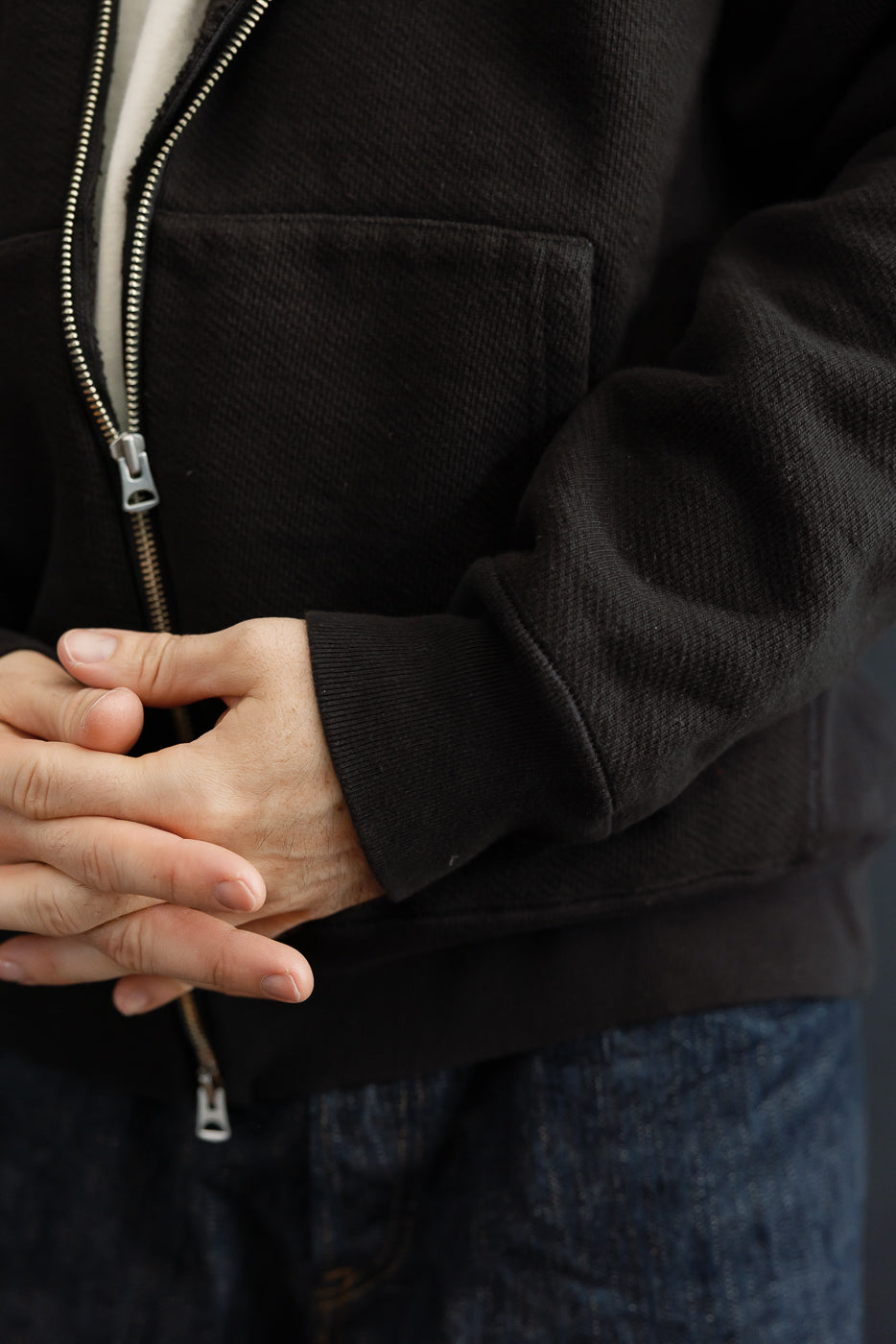Close-up of the front of a male model's black zip-up hoodie, showing textured fabric and hands clasped.