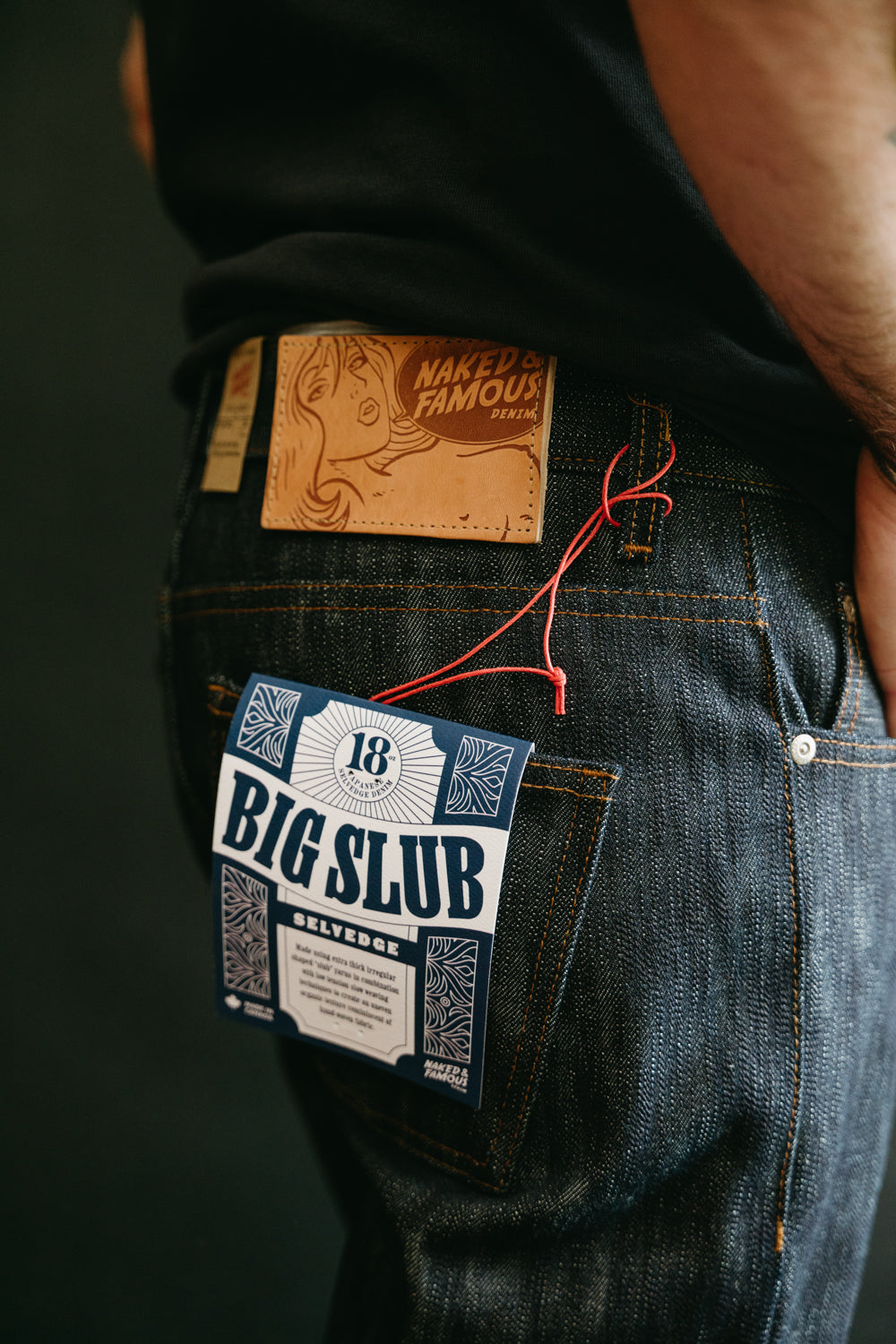 Close-up of male model’s back right side wearing Naked & Famous 18oz Big Slub Selvedge jeans, showing leather patch, rear pocket, and product tag. Model is 5'9