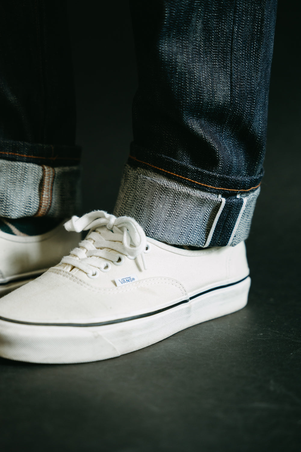 Close-up of male model’s lower leg in Naked & Famous 18oz Big Slub Selvedge jeans, front view cuffed hem showing selvedge detail, paired with white Vans sneakers. Model is 5'9