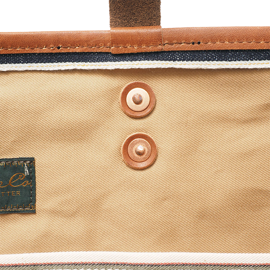 Close-up of tote bag interior showing beige twill fabric, leather trim, rivet buttons, and inner tag.