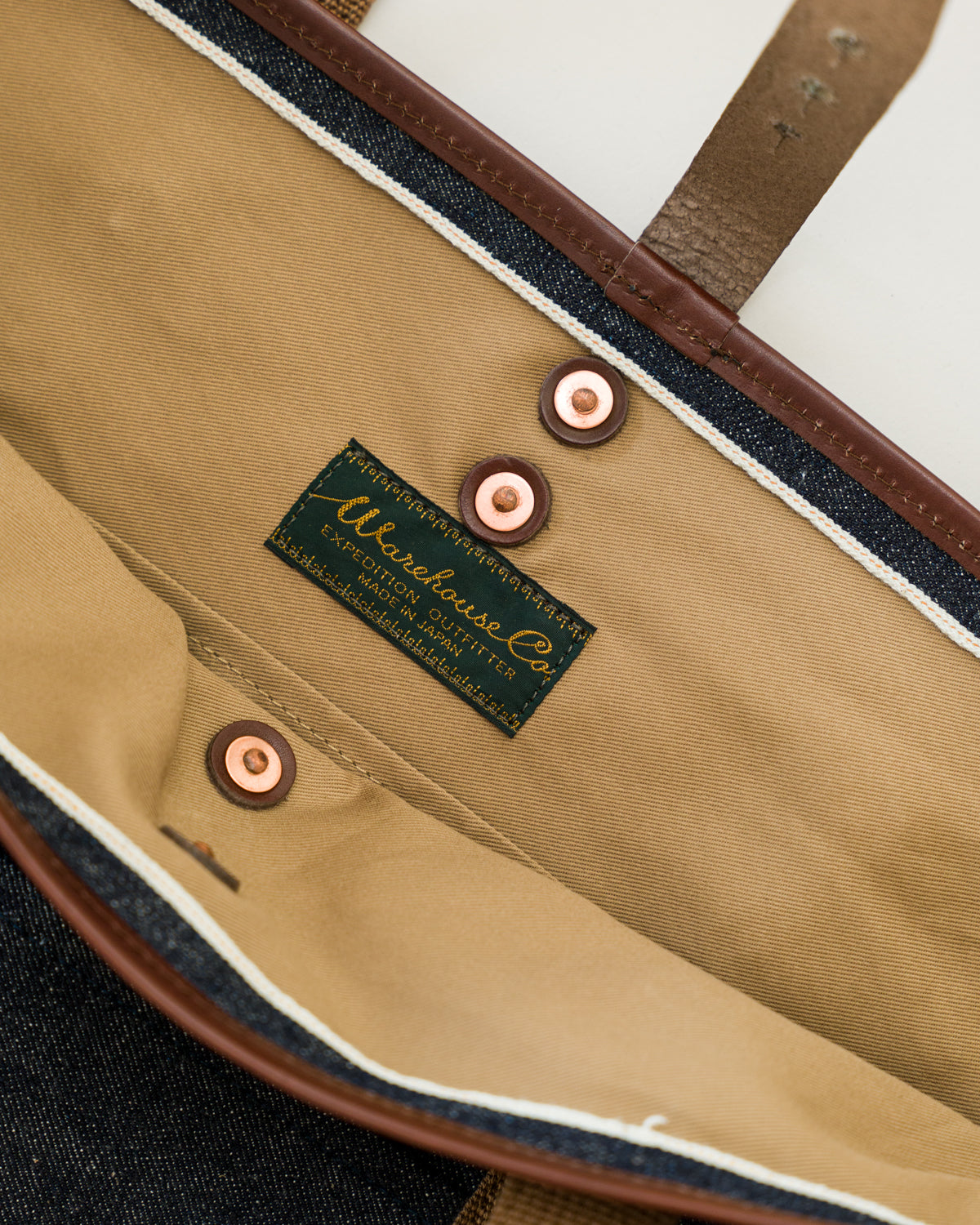Close-up of tote bag interior showing tan lining, copper rivet, label, and leather trim.
